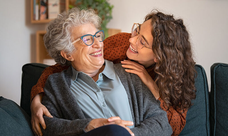 Young woman smiling and hugging her grandmother from behind.